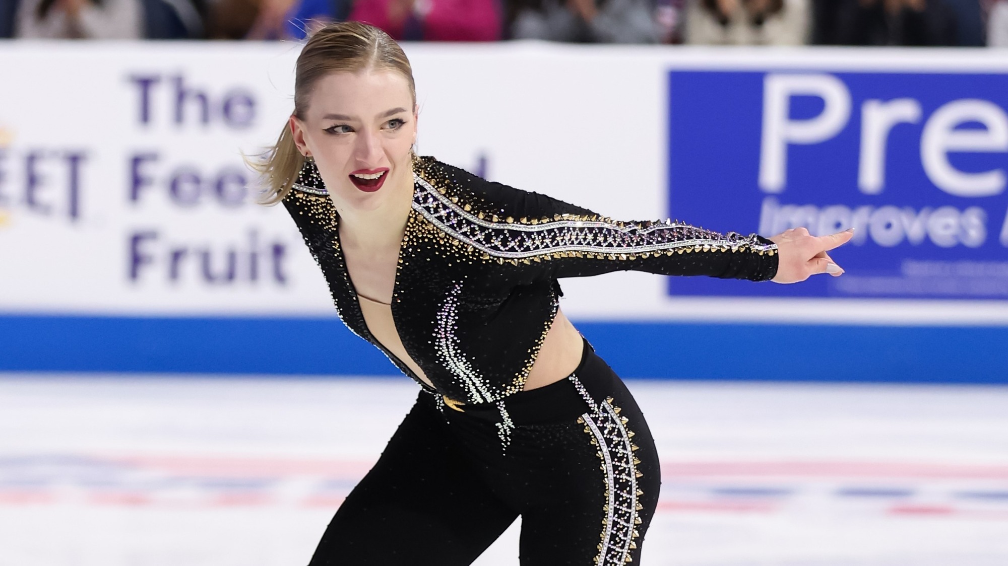 Amber Glenn skates toward the camera, her hair pulled back in a pony tail. The picture is close up, taken from her thigh up. She wears a black bodysuit with silver and gold jewels.