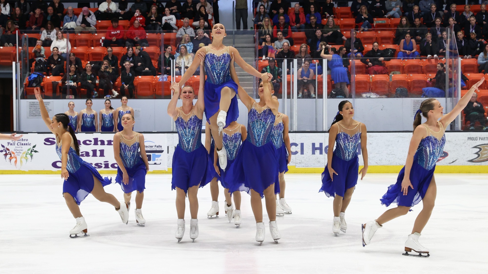 Miami University competes in their free skate. The team is wearing matching blue and purple skating costumes. In the middle, two skaters hold a teammate up in the air. 