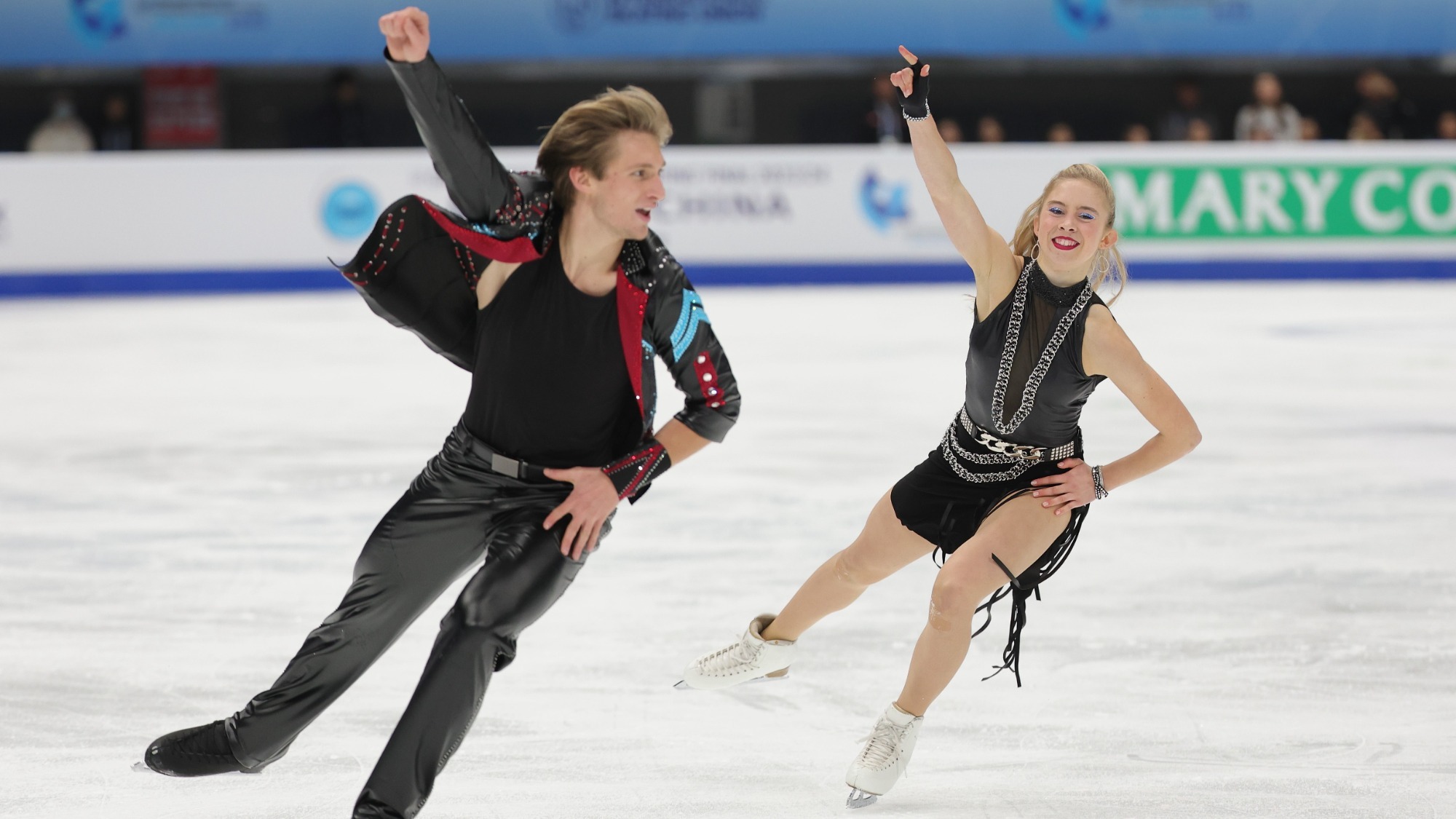 Artem Markelov and Leah Neset skate toward the camera during their rhythm dance. They have the same movement, their right arm up in the air and left arm on their hip. She wears a leather dress with big bold silver jewelry. He wears black leather pants and a black shirt with a black lather jacket that has red and light blue detail. 