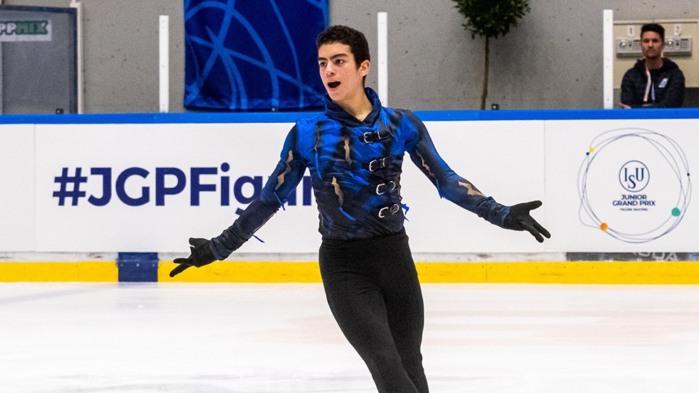Jacob Sanchez, with his arms out to the side while he skates, an excited look on his face. Taken from the thigh up, he wears black pants and a blue and black top.