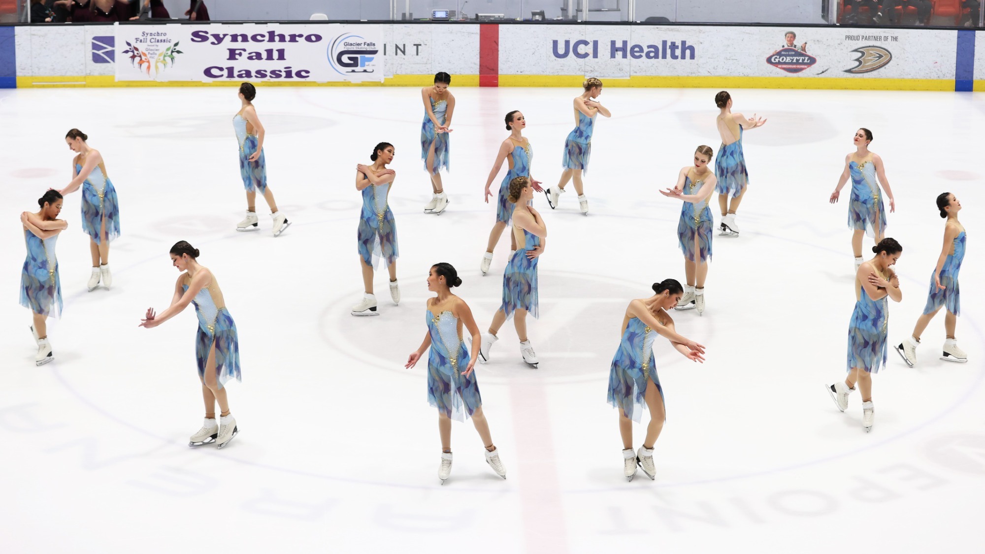 The Lexettes hit their starting pose, spread out across the ice in a circle, for their short program. They wear dresses with different blues.