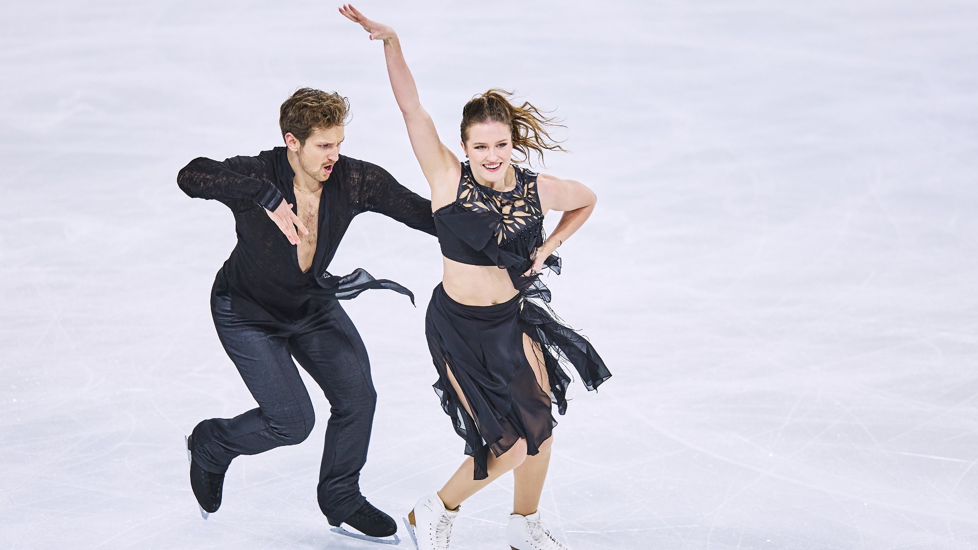 Christina Carreira and Anthony Ponomarenko on the ice. Both wear black. He skates behind her, her holding her waist. She holds his hand on her waist, her other arm up in the air.