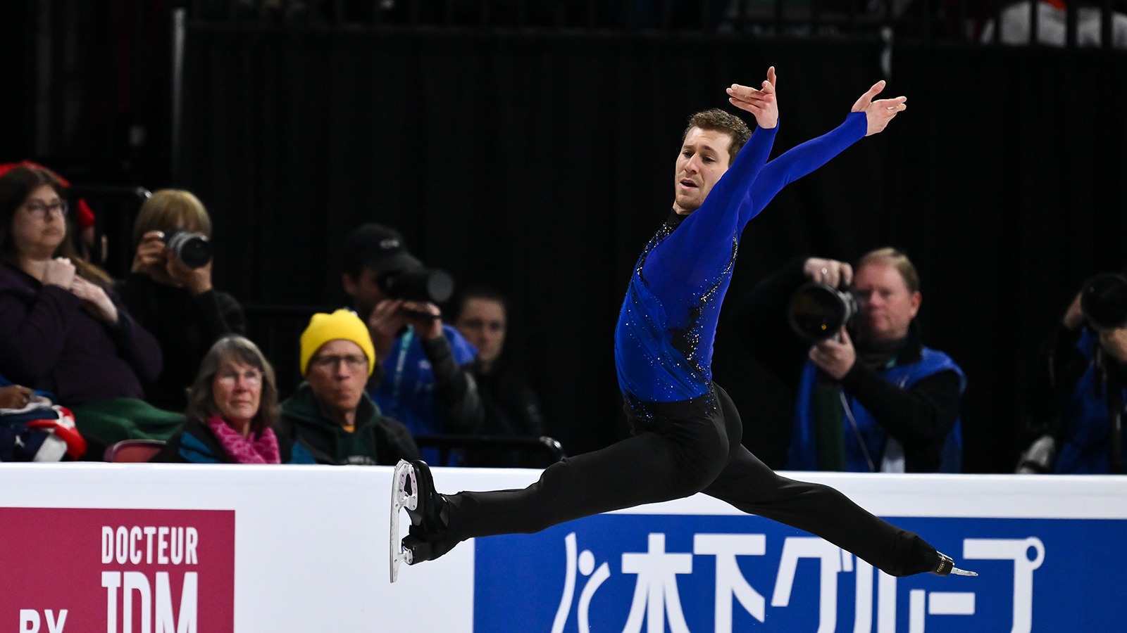 Jason Brown, dressed in a blue shirt and black pants, soars through the air, legs and arms extended.