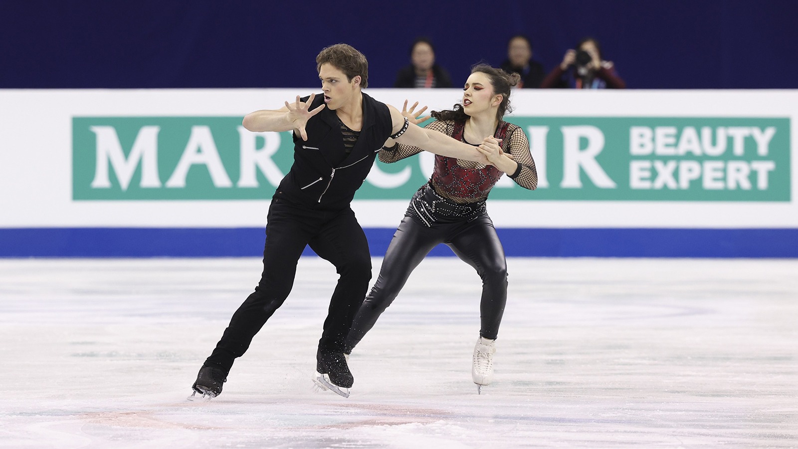 Michael Parsons, dressed in all black, and Caroline Green, dressed in black leather pants, a fishnet top with a multi-colored designed undershirt, skate side by side, showing their chemistry and unison.