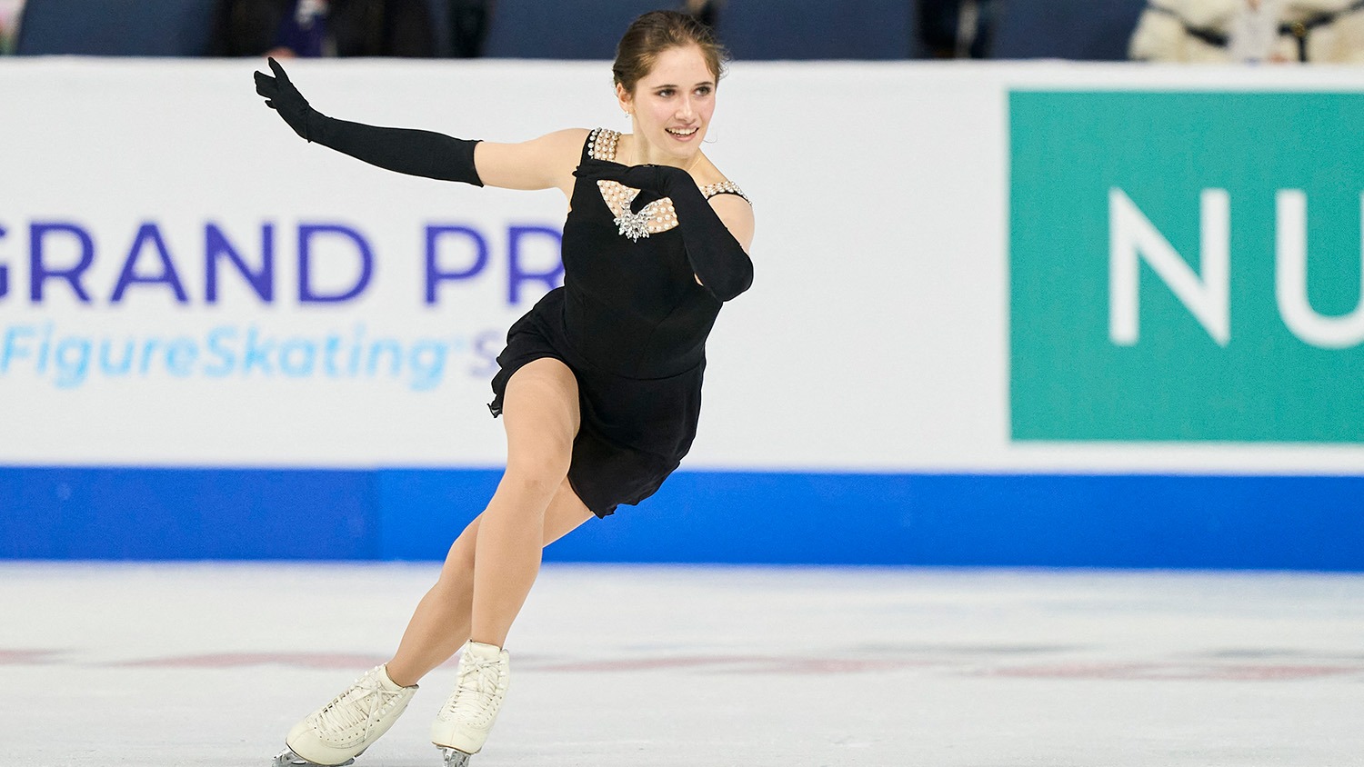 Isabeau Levito, wearing a black dress with pearl embellishment at the top and black gloves, skates to the right, a smile on her face.