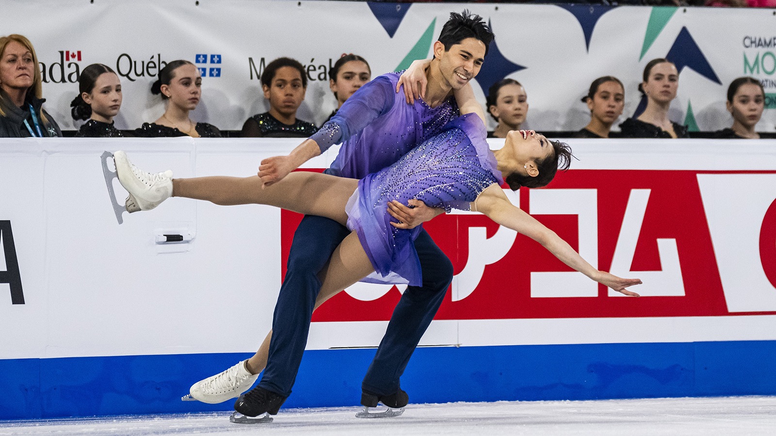 Emily Chan and Spencer Howe perform on the ice with Spener holding up Emily by the waist. Emily is a young woman with short black hair wearing a purple skating costume. Spencer is a young man with short black hair wearing a matching long sleeve top and black pants