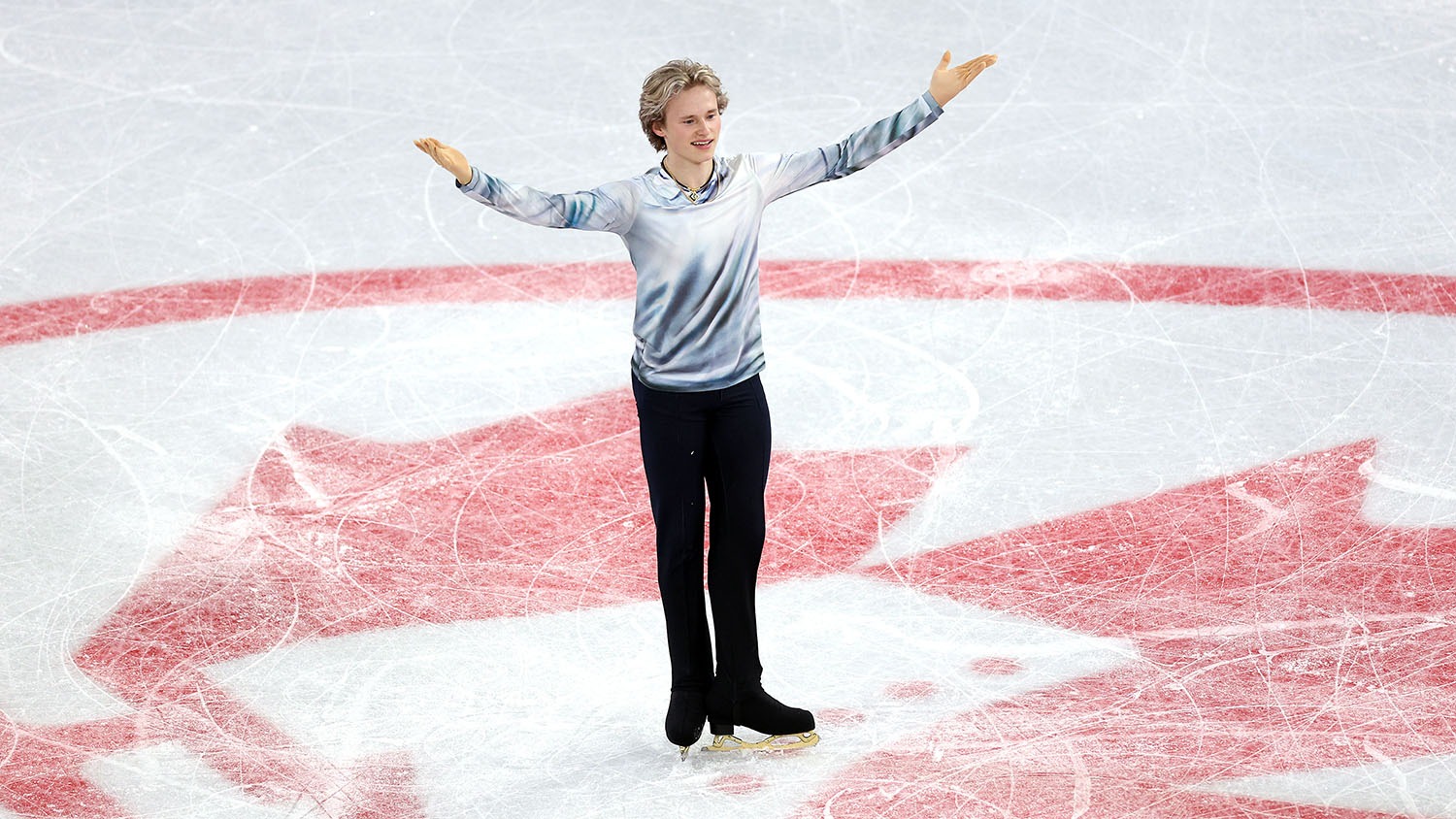 Ilia Malinin salutes the crowd at center ice, smiling. He stands over the Skate Canada logo, and wears black pants and skates as well as a grey top.