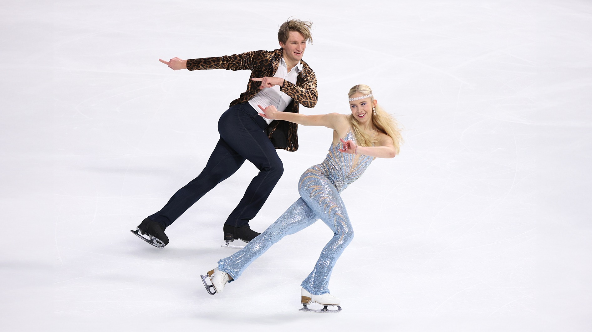 Leah Neset and Artem Markelov skate to the right while pointing to the left with both hands. Leah is wearing a 70s inspired outfit with a flower headband and Artem is wearing a leopard print jacket