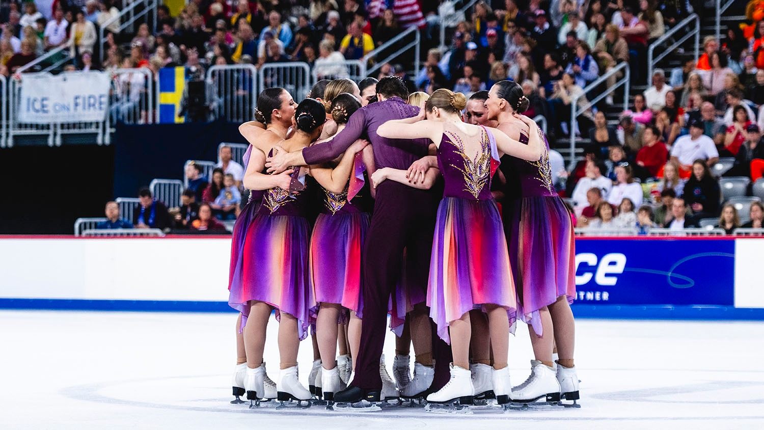 The Haydenettes huddle together in a circle on the ice after their skate, their arms wrapped around each other. The crowd cheers in the background.  They dresses iwith purple tops and gold details while the shirt includes purple, red, orange and yellow.