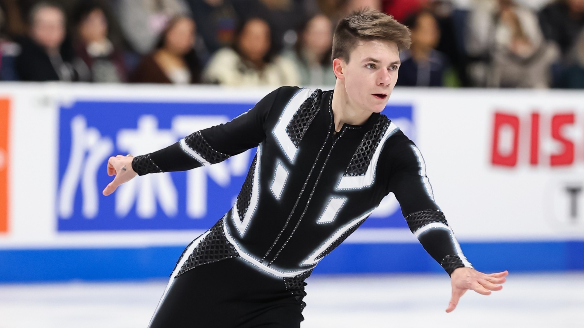 Taken from the waist up, Maxim Naumov looks forward with his arms outstretched. He is a young man with short brown hair wearing a black long sleeve body suit with white stripes to match his Tron-inspired program. 