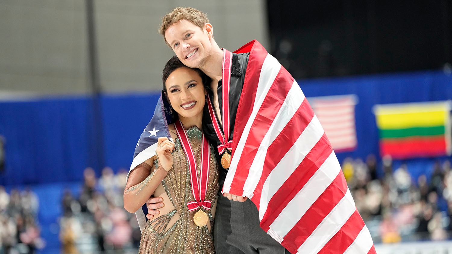 Gold medalists Madison Chock and Evan Bates of the United States side hug as they skate during the victory ceremony at NHK Trophy. They both wear gold medals, the U.S. flag draped around them as they smile.