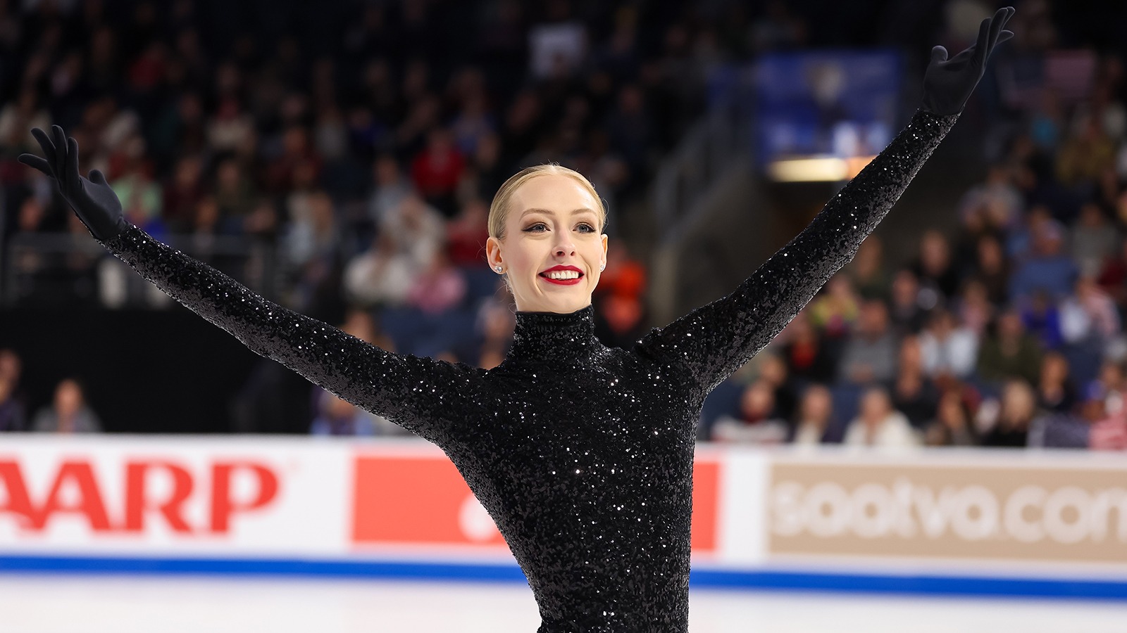 Bradie Tennell smiles and salutes the crowd at 2024 Skate America. Bradie is a young woman with blonde hair tied back in a bun. She is wearing a long sleeve black and sparkly skating costume with a turtleneck neckline and attached gloves.