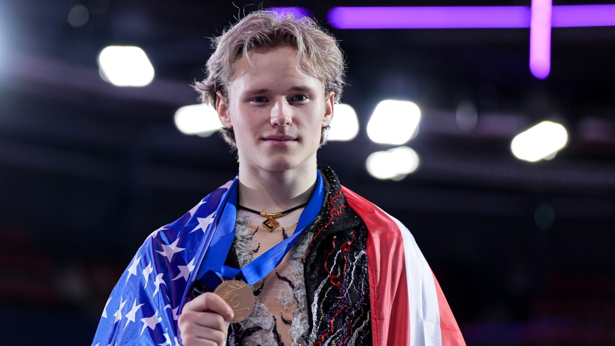 Ilia Malinin smiles at the camera while holding his Grand Prix Final gold medal in his right hand. A United States flag is draped around his shoulders. Ilia is a young man with blonde hair wearing a black skating costume with red and white detailing around the v-neck neckline. 