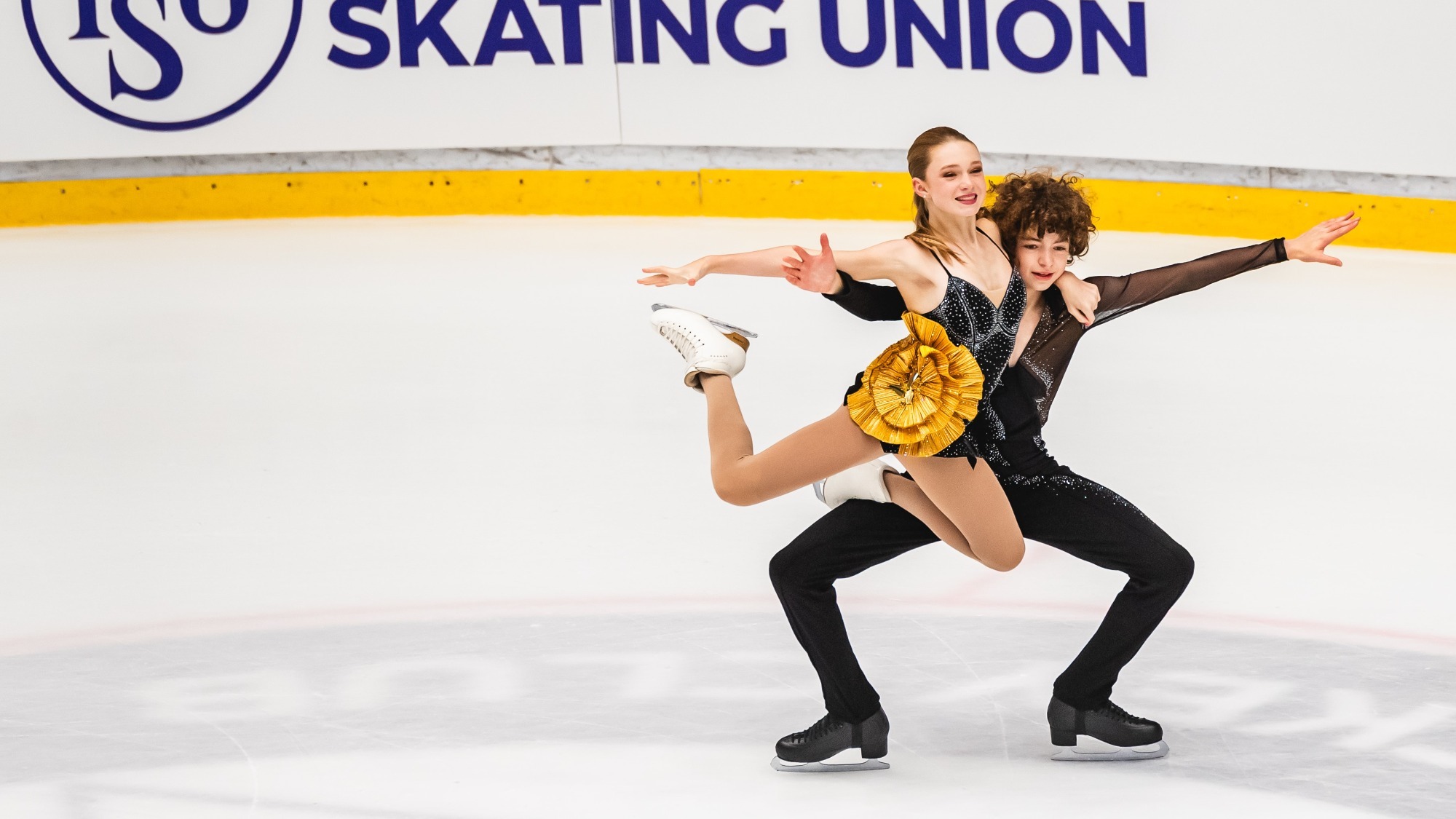 Olivia Ilin and Dylan Cain compete at JGP Hungary. Ilin is a young woman with long blonde hair and Cain is a young man with curly brown hair. Cain holds up Ilin in a lift. Cain is wearing a long sleeve black top with black pants and Ilin is wearing a black skating costume with a gold bow detail at the hip