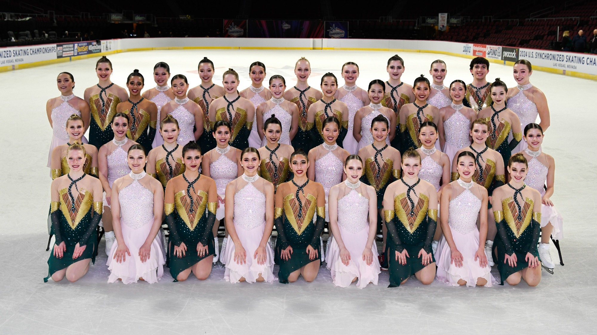The 2024 World Junior Championships Team: Teams Elite and Skyliners. They pose for a formal photo on the ice in four rows alternating between Teams Elite and Skyliners athletes. The Skyliners wear light pink dresses while Teams Elite wears gold and green dresses with a snake detail around the neck.