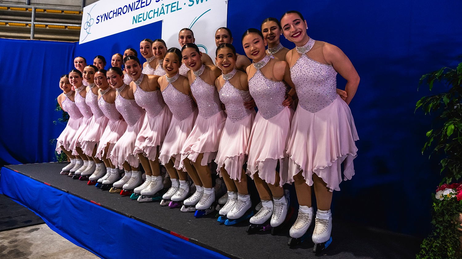 Skyliners junior, in light pink dresses, poses for a photo in the kiss and cry. The first row bends their knees, the second row standing behind.