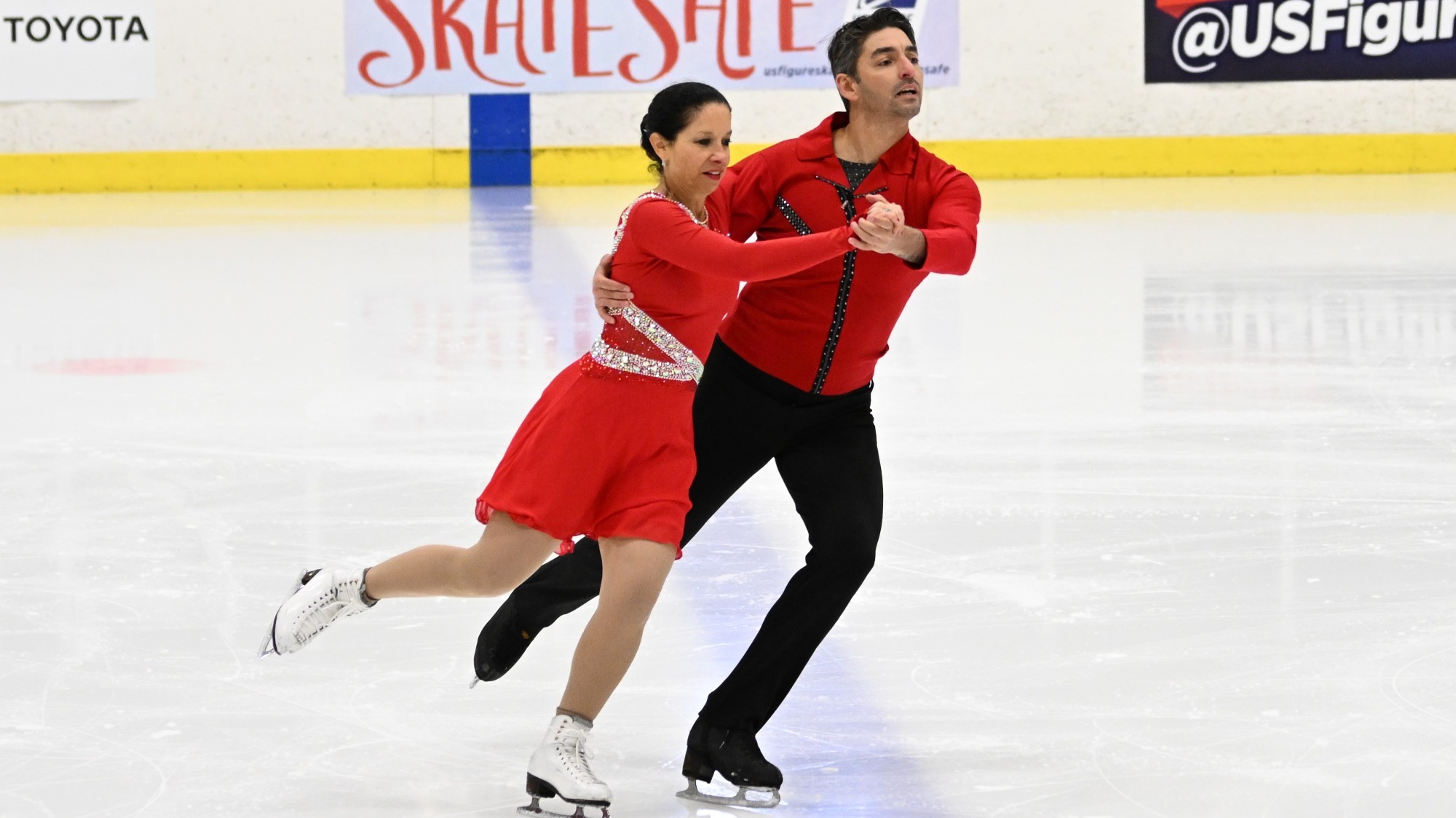 A man and a woman skate holding on to each other wearing coordinated red costumes. The woman is wearing a red long sleeve skating dress with a silver belt, and the man is wearing a red button-down with black pants