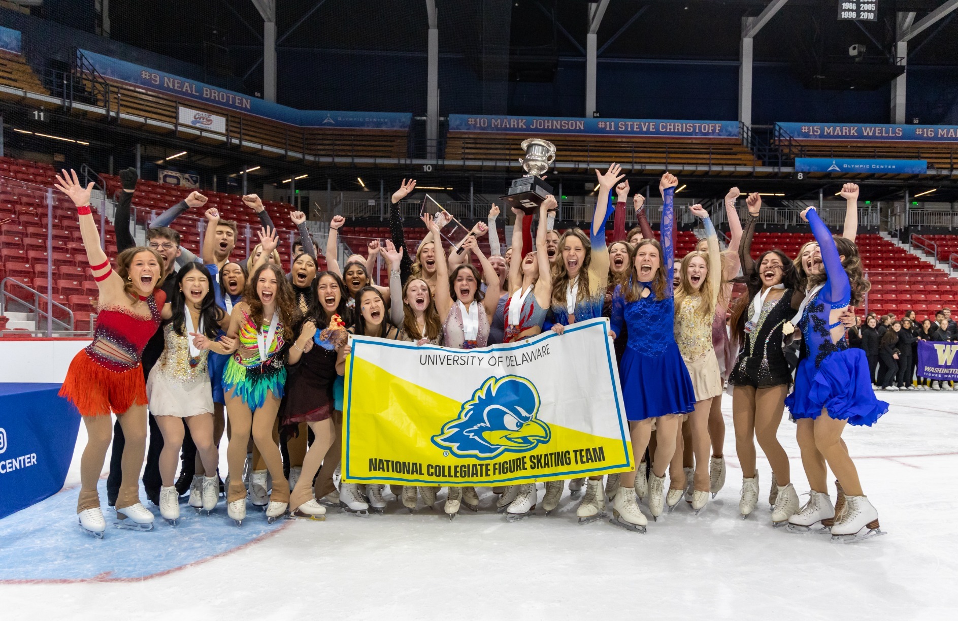 The University of Delaware Figure Skating Team cheers while one skater holds up the National Intercollegiate Final trophy after they just won. They stand behind a large University of Delaware National Collegiate Figure Skating Team flag that is yellow and white with the blue hen logo in the center