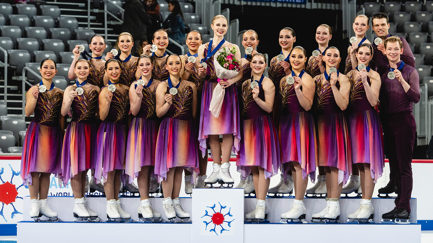 The Haydenettes stand on the podium, each athlete holding up their silver medal. They wear their free skate costumes, which are purple with red and orange hues. 