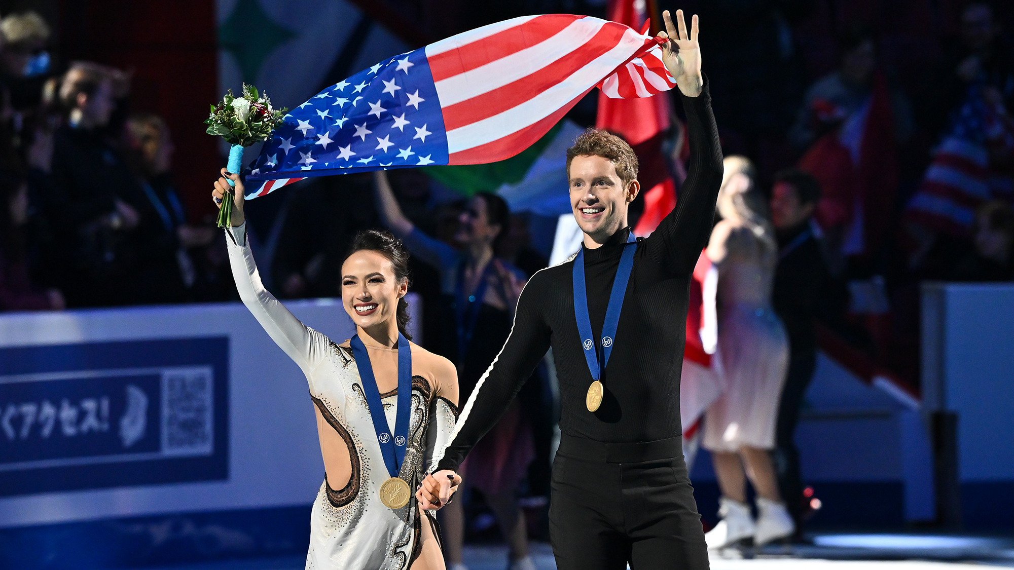 Madison chck and Evan bates hold hands, gold medals around their necks. With their outside arm, they wave to the crowd, holding the U.S. flag in the air behind them.