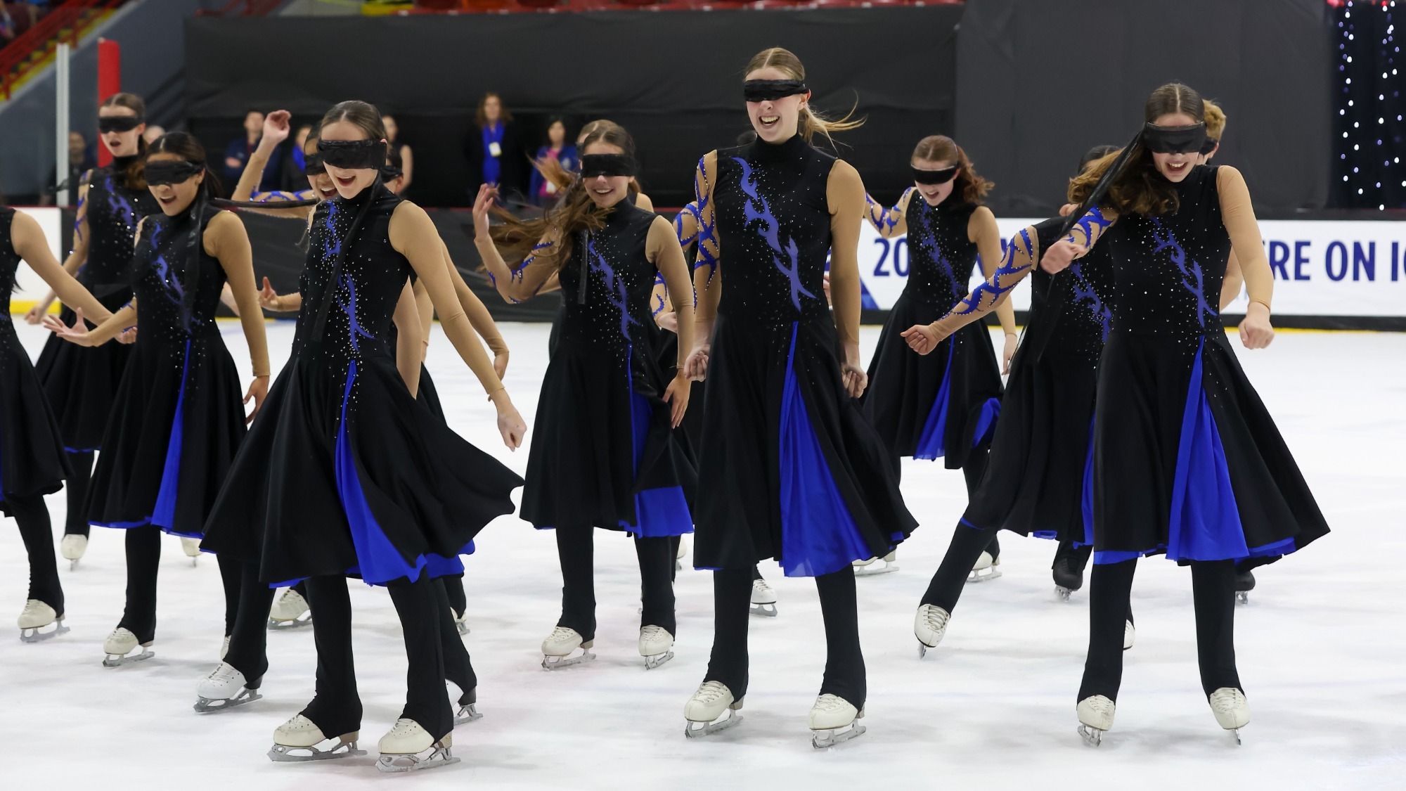 Act 1 of Boston stands on the ice facing forward. Each skater is wearing a long black skating costume with a royal blue underskirt and flames across their chests in the same color. Each skater is wearing a black blindfold over their eyes. 