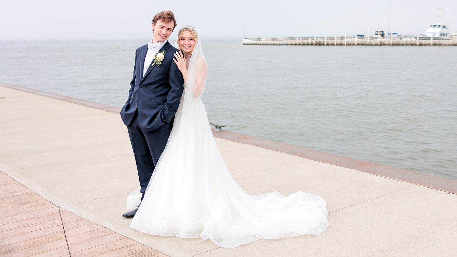Logan Bye (left) with Eva Page (right). They stand on a dock in front of a lake and marina. He wears a navy tux while she wears a wedding dress and veil. Both smile for the camera.