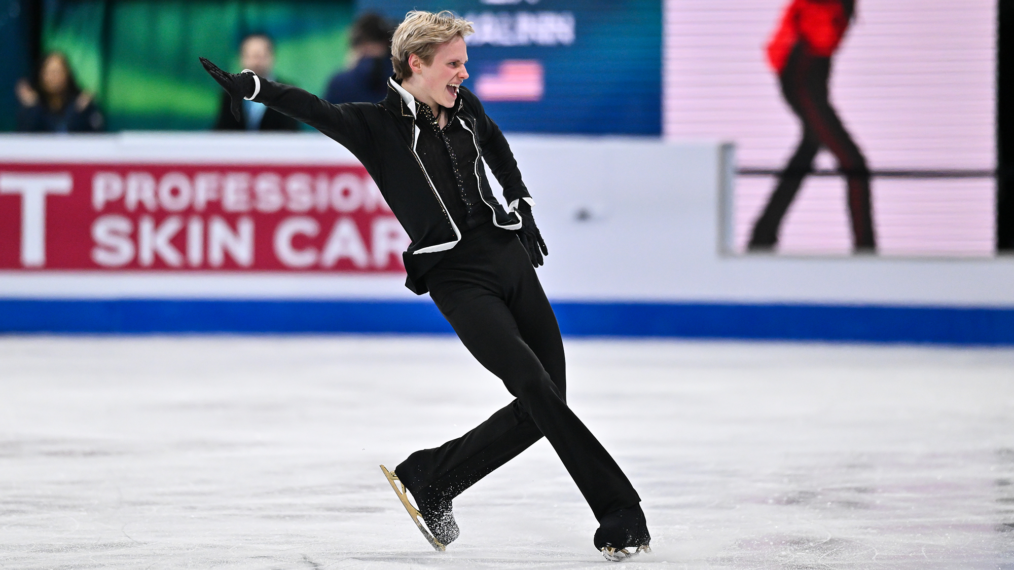 Ilia Malinin, in a black shirt, jacket, pants and gloves, looks to the right, his left hand out, a fierce expression on his face.