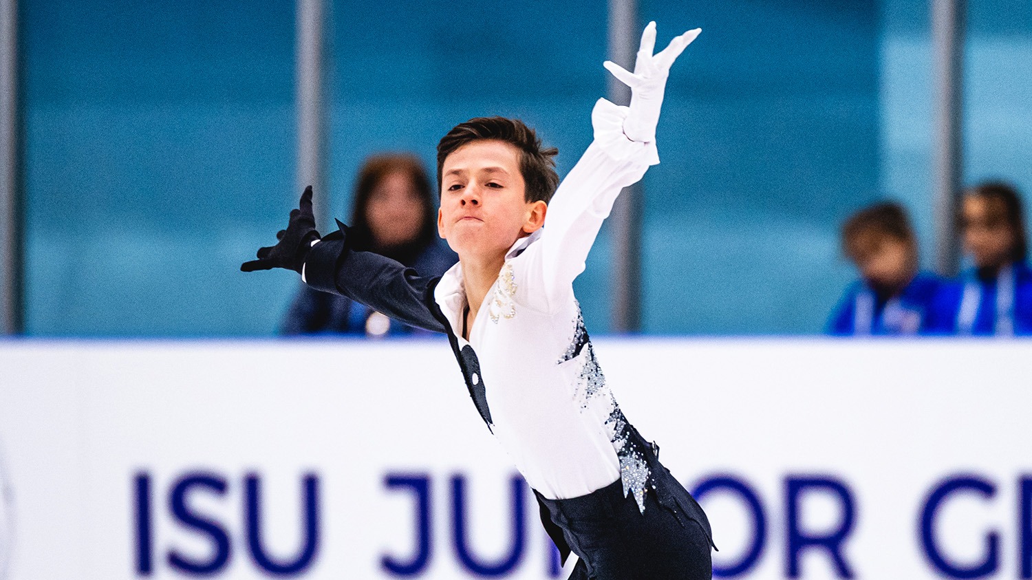 Aleksandr Fegan, in a picture from the waist up, skates to the left with his arms out wide, a serious look on his face. He wears black pants and a white and black top. 