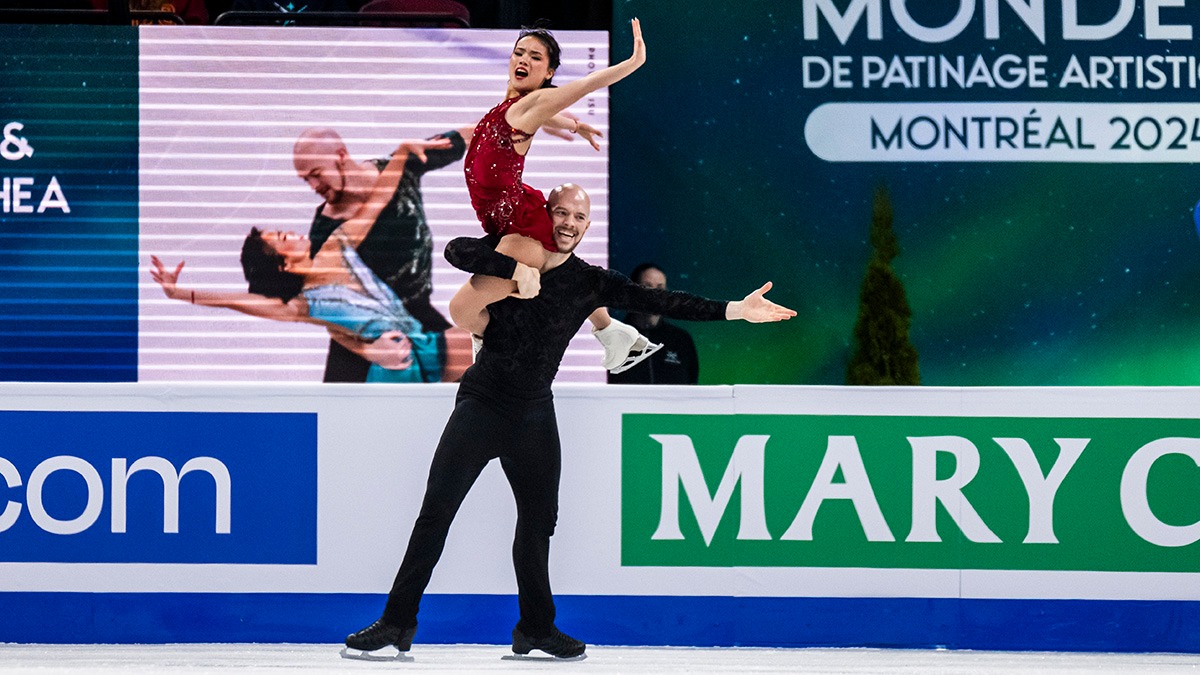 Ellie Kam and Danny O'Shea perform a lift on the ice. She sits on his shoulder as he holds her with one hand. Danny, wearing all black, smiles, while Ellie, in a red dress, sits up tall.