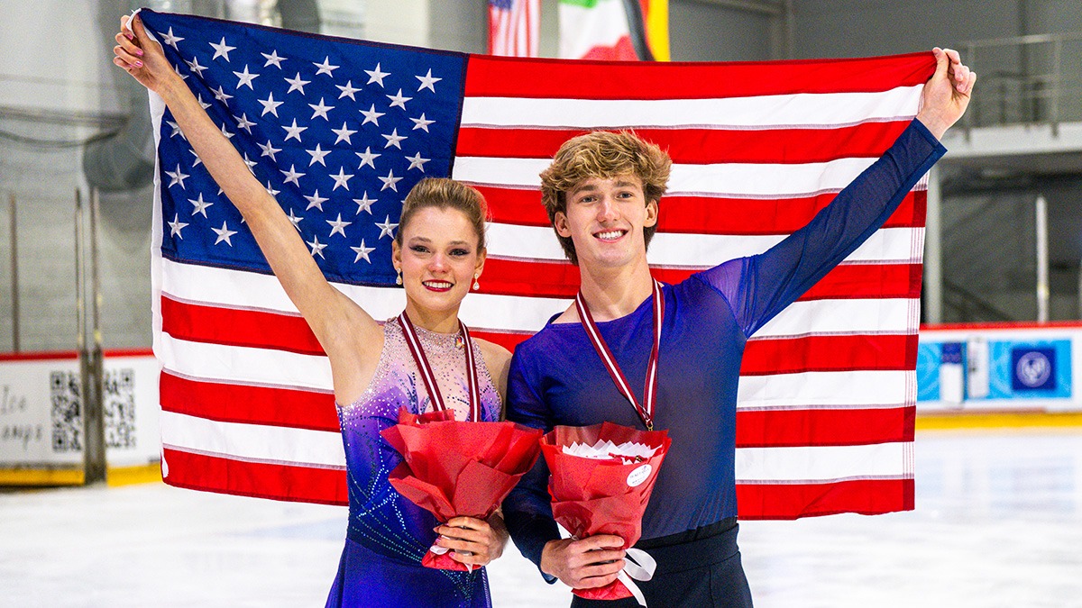 Caroline Mullen and Brendan Mullen hold up the U.S. flag behind them, holding flowers in their other hand, silver medals around their necks. The ice rink is in the background. They both wear their blue free skate costumes.