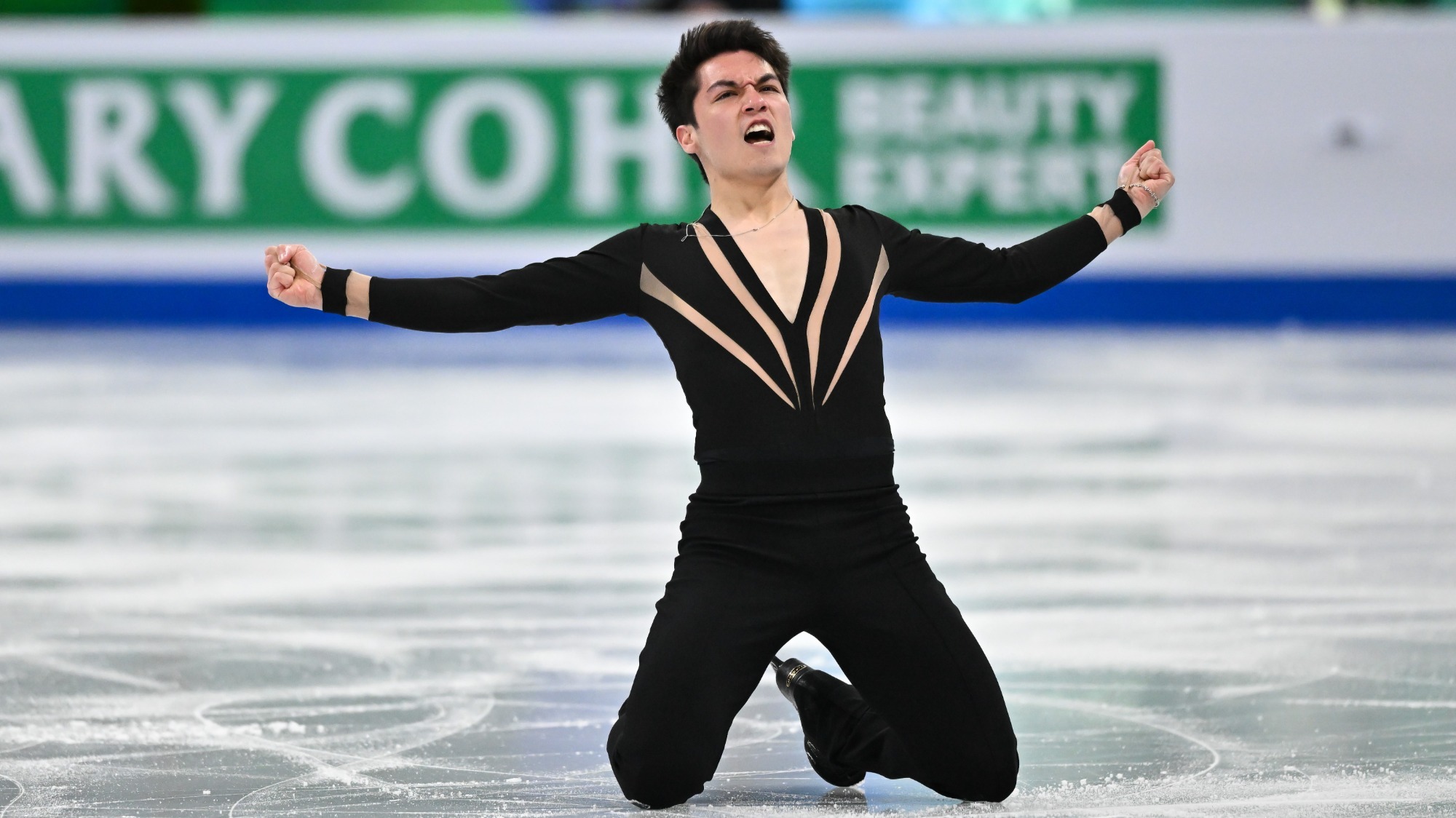 Camden Pulkinen kneels on the ice with his arms outstretched, looking triumphant. He is wearing a black top with gold details along the neckline and black pants. 