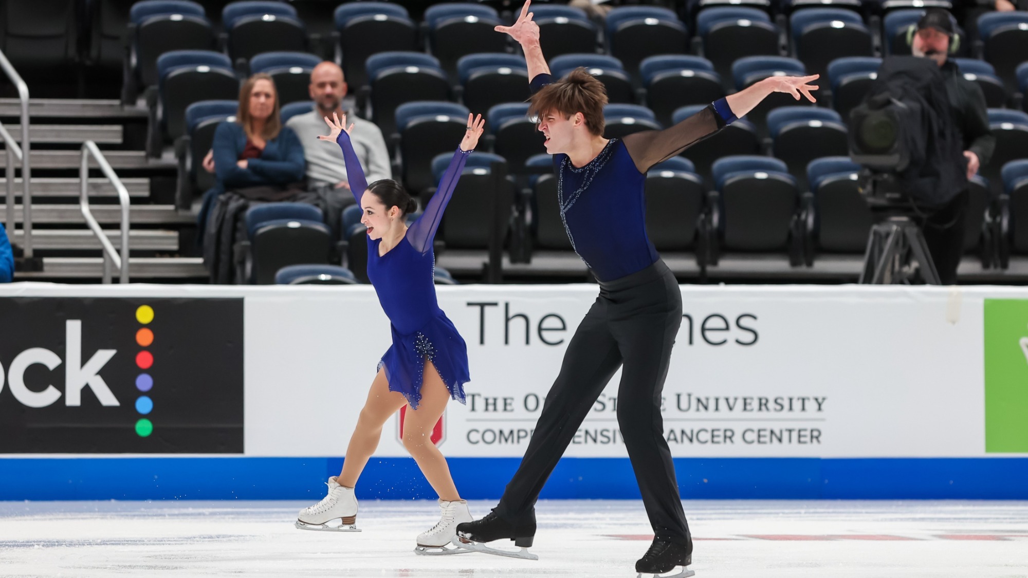 Katie McBeath and Daniil Parkman skate to the left with their arms extended upward. They both have a determined look on their face. Katie (left) is wearing a long sleeve blue skating costume and Daniil is wearing a long sleeve black shirt and black pants. 