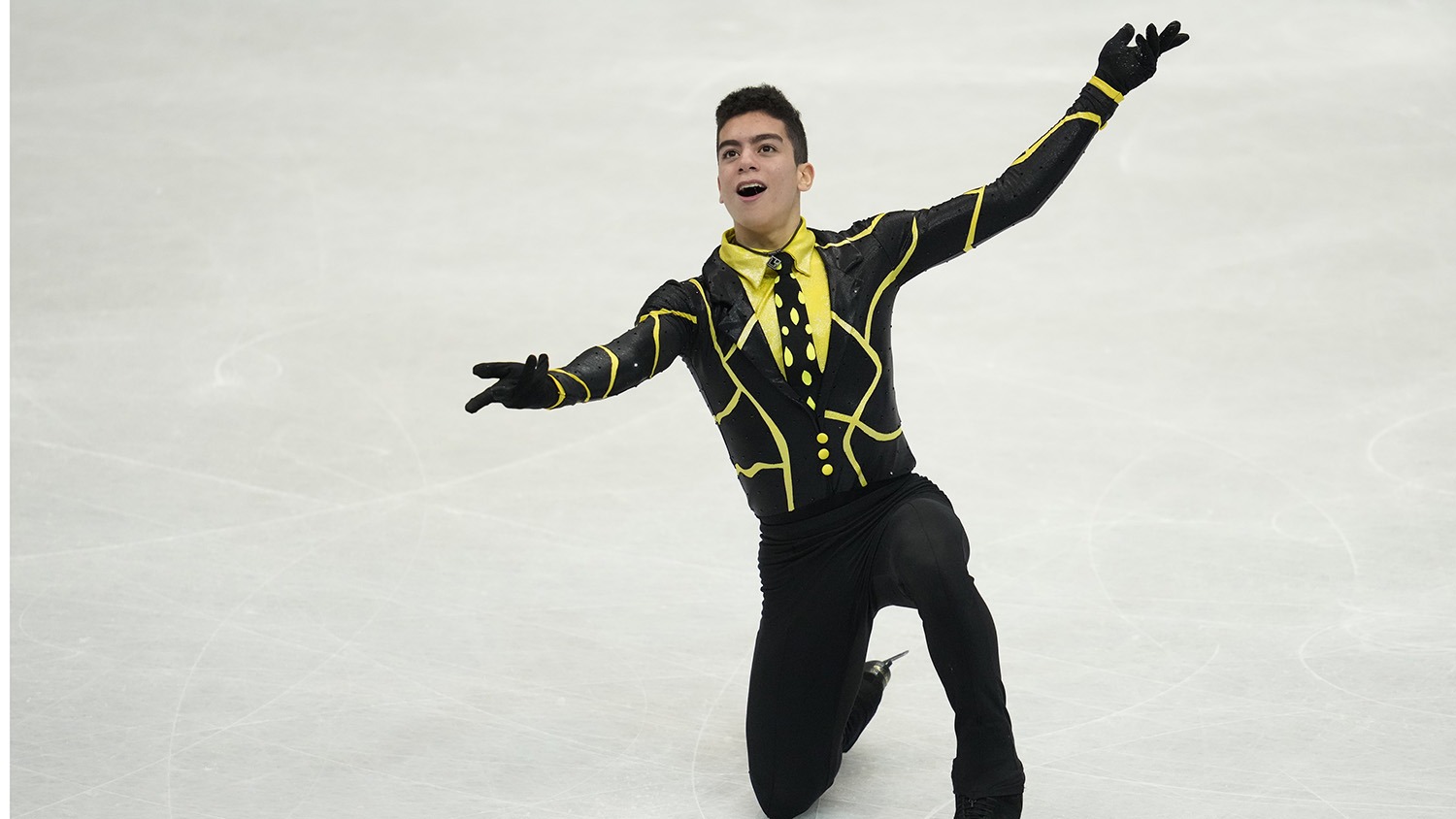 Jacob Sanchez, wearing a black and yellow top and black pants, kneels on the ice, a fun expression on his face and his arms out to the side
