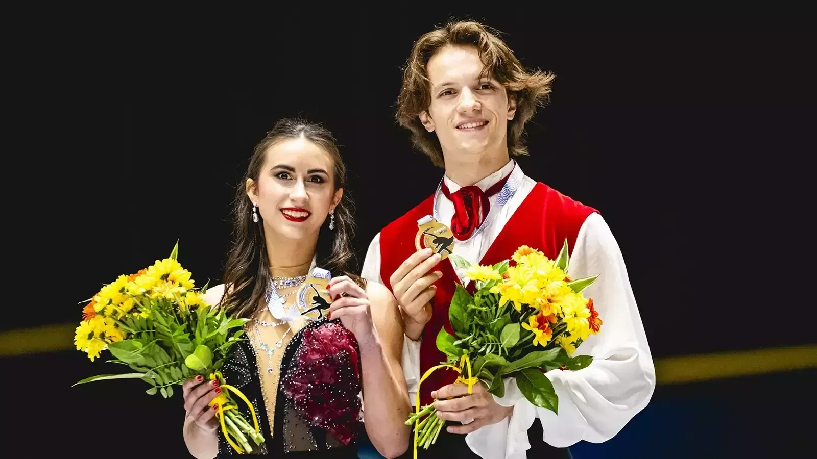 Katarina Wolfkostin and Dimitry Tsarevski, holding yellow flowers, display their gold medals on the podium.