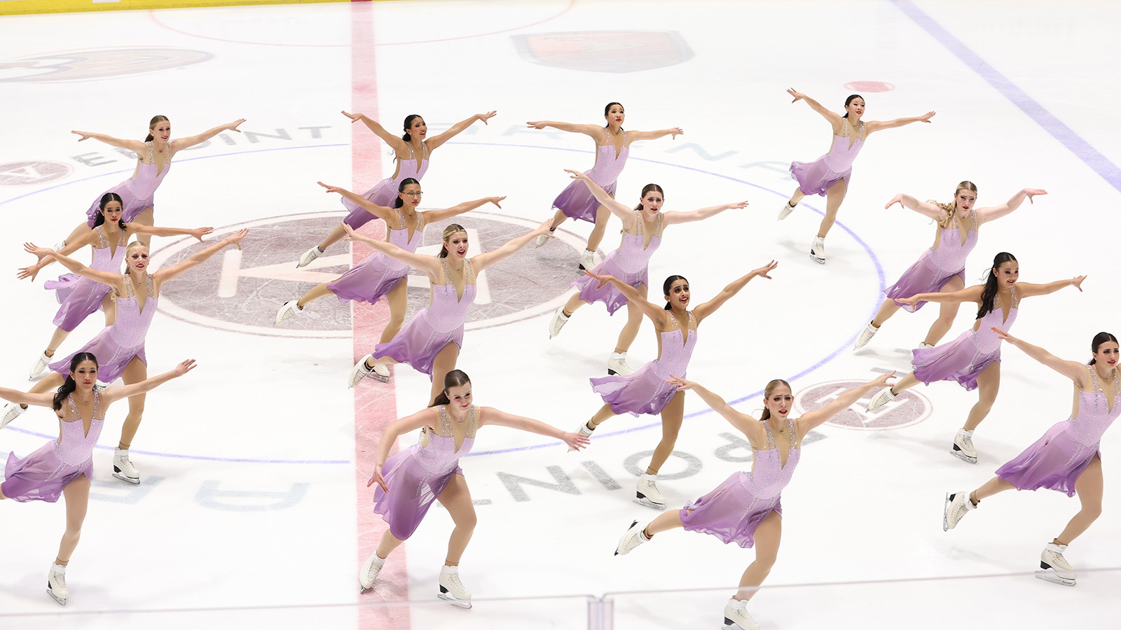 The Lexettes skate in four parallel lines. Each skater has their arms outstretched in unison. They are wearing matching purple skating costumes