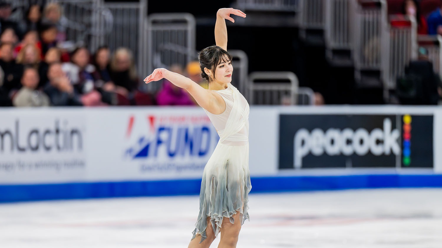 Alysa Liu, in a white dress that fades to grey stands on the ice, her arms above her head and pointed out, at the U.S. Championships.