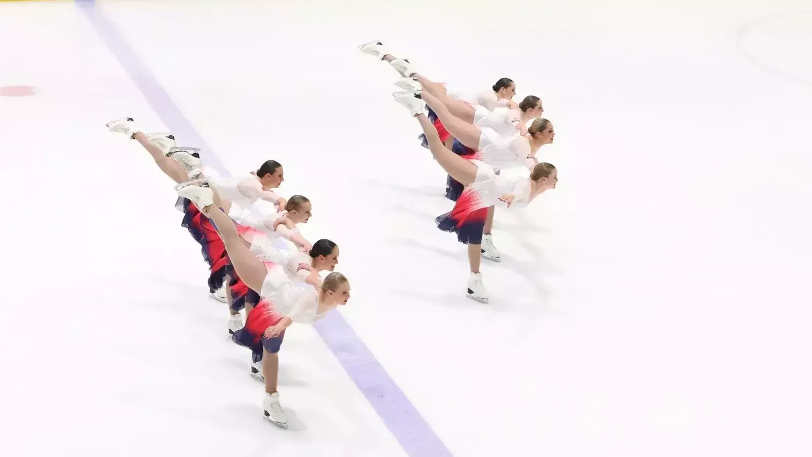 The Haydenettes skate together in two symmetrical lines. Each skater in leaning forward with their left legs extended in the air behind them. They are all wearing matching white skating costumes with pink and black ombre skirts