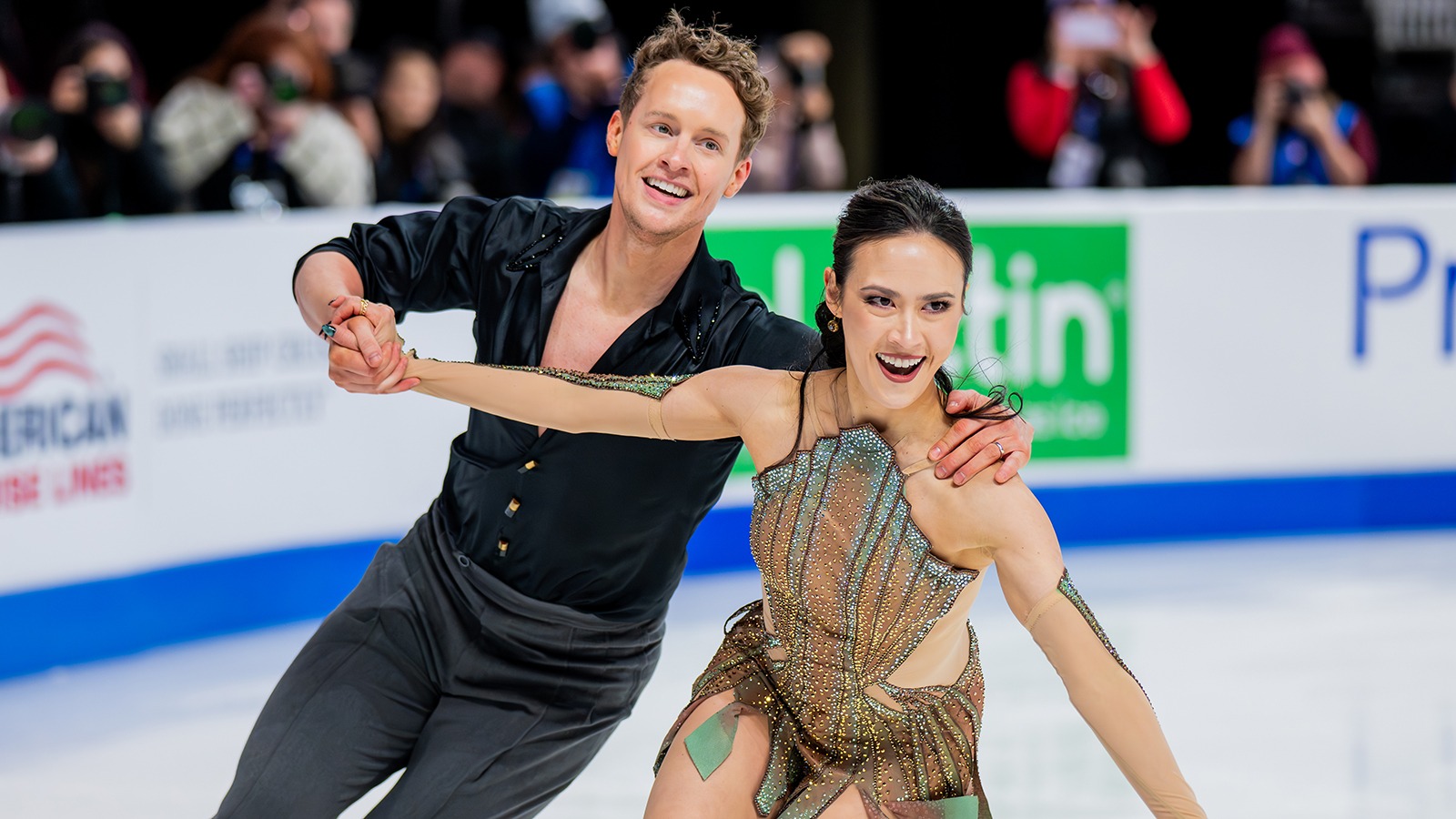 Madison Chock and Evan Bates skate hand in hand. Madison is a young woman with long black hair wearing a copper and green dress inspired by the statue of liberty and Evan is a young man with short curly blonde hair wearing a button down black shirt with the sleeves rolled up to the elbows. 