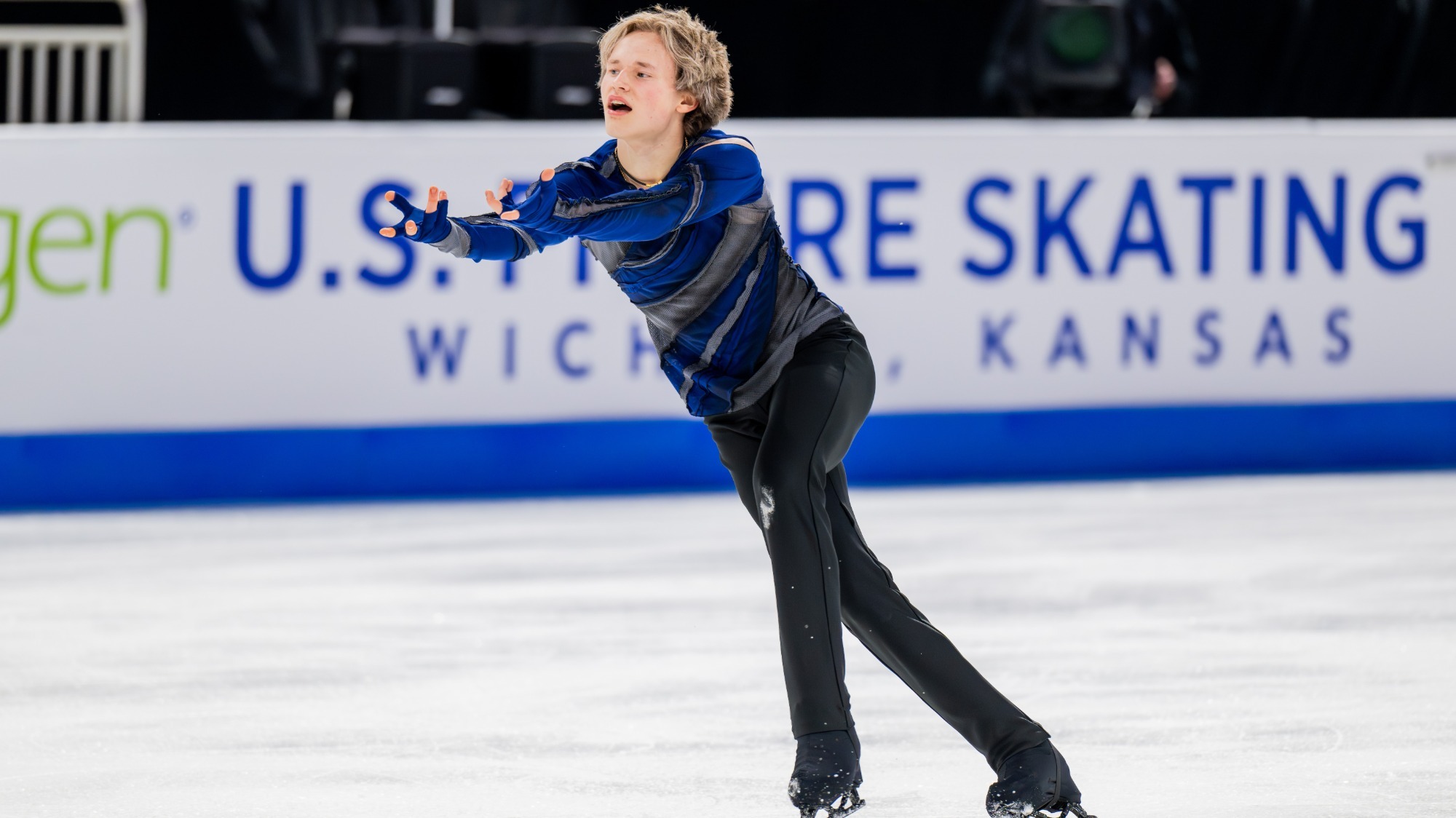 Ilia Malinin skates forward with his arms outstretched in front of him. He is a young man with short blonde hair wearing a dark blue long sleeve shirt with black stripes and black pants. 
