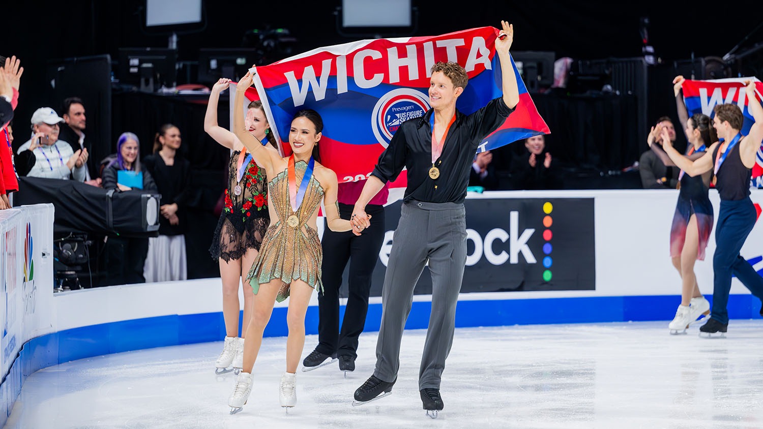 Madison Chock and Evan Bates skate by the crowd during the victory ceremony, smiling and holding a Wichita flag behind them