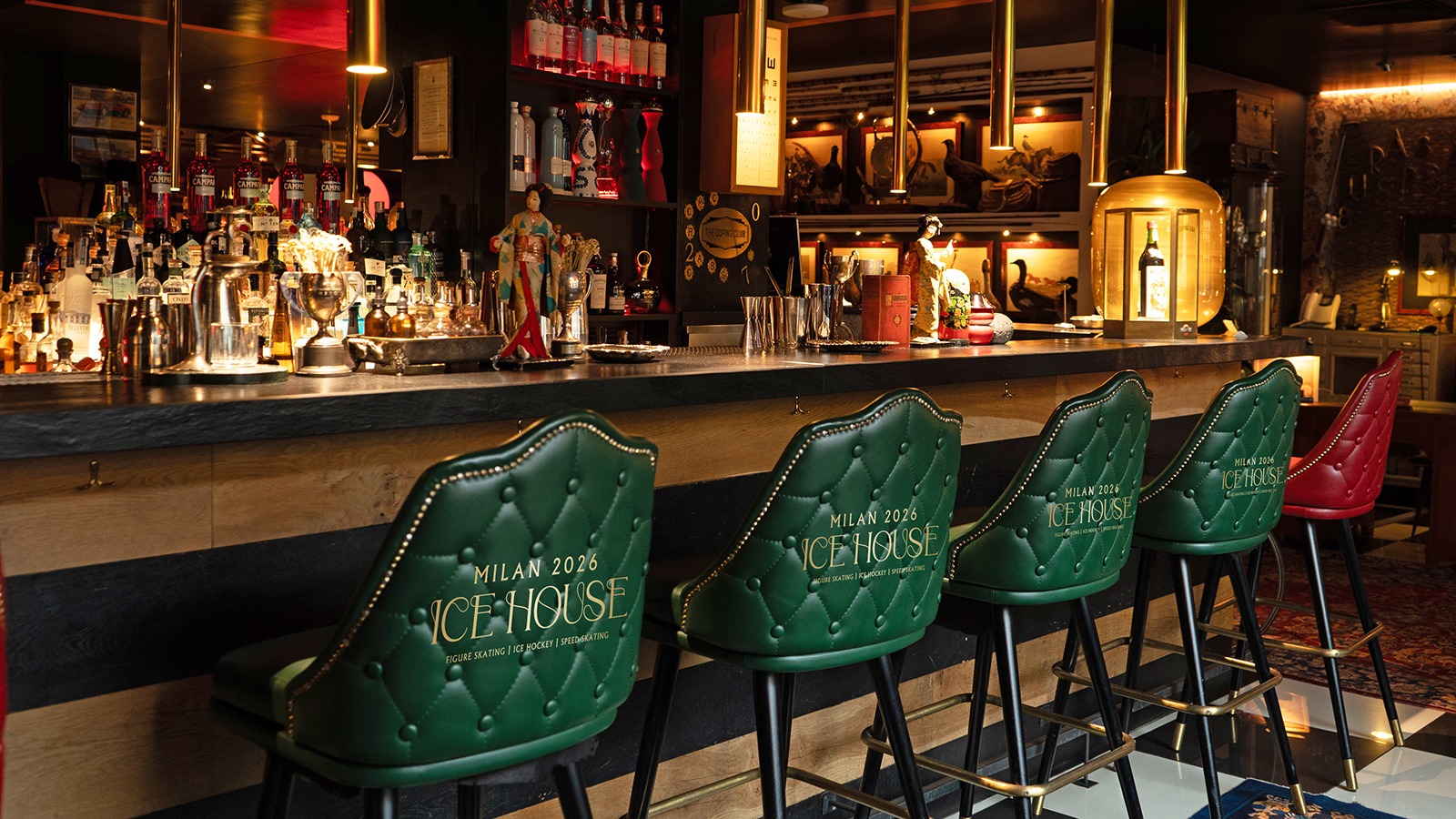 Photo of a bar with green bar stools that say Milan 2026 Ice House. Below that it says figure skating, ice hockey, speedskating Behind the bar are several bottles on the shelves. 