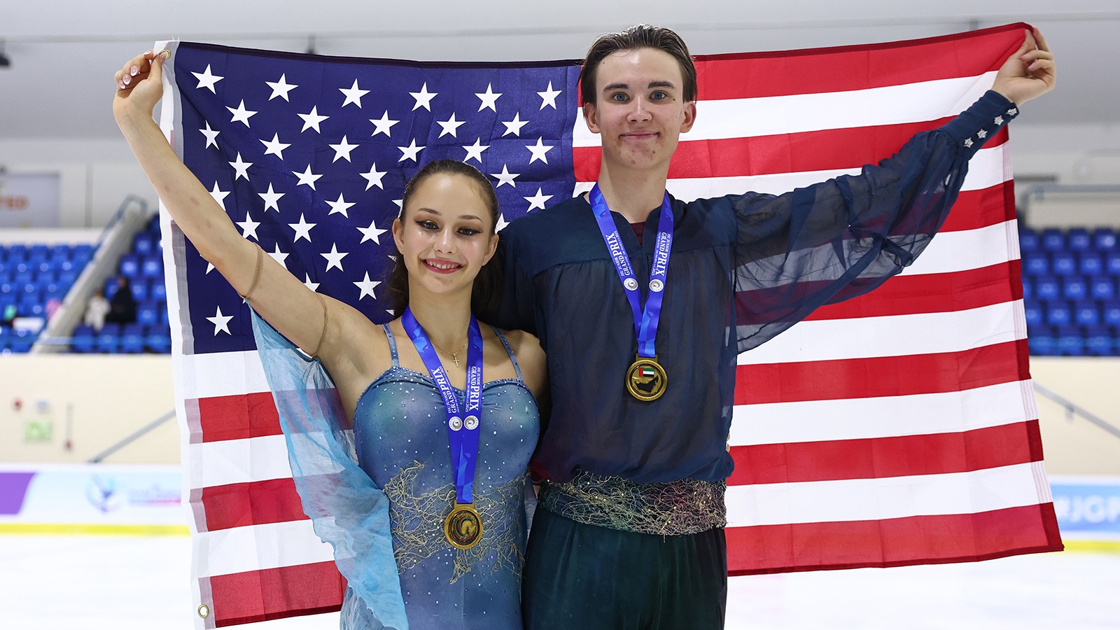 Hana Maria Aboian and Daniil Veselukhin stand smiling with gold medals around their necks. They are both holding the corners of an American flag behind them. Hana (left) is a young woman with long brown hair tied back in a ponytail wearing a blue skating costume with spaghetti straps and blue fabric resembling wings down the side. Daniil (right) is a tall young man with short brown hair wearing a dark blue long sleeve top with sheer sleeves and a beaded belt. 