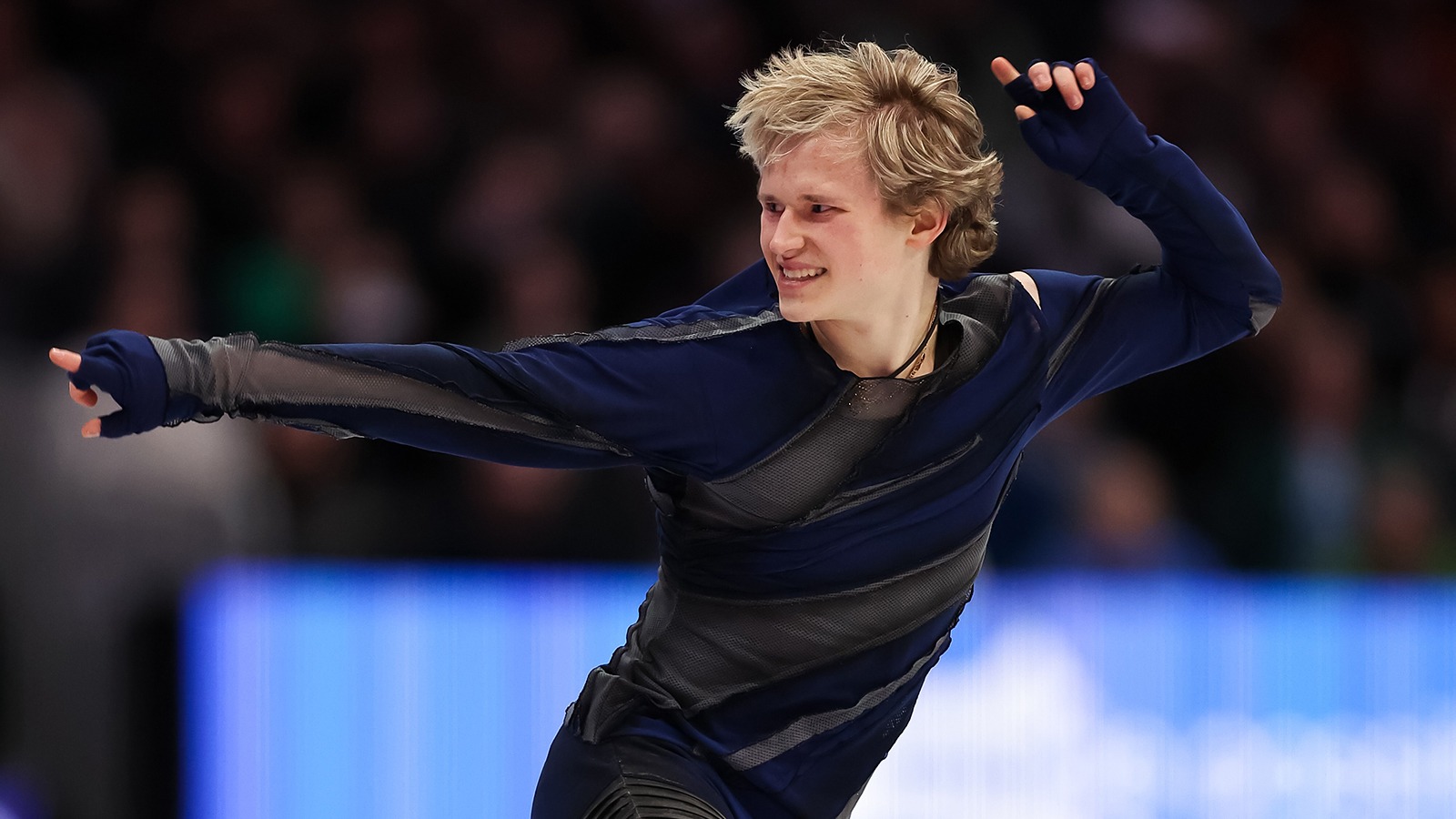 Ilia Malinin, dressed in dark blue and dark gray, performs at the World Championships. He is smiling, pointing with one arm extended out and one arm extended up.