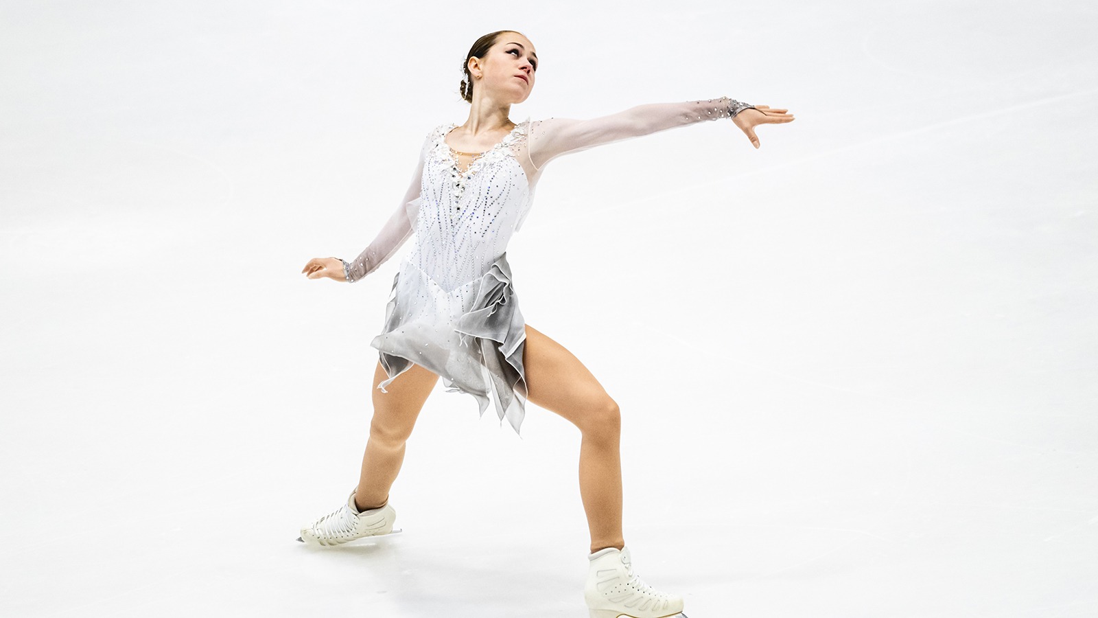 Sophie Joline von Felten, dressed in all white, performs a dramatic program at JGP Poland. Her back is arched and her arms are extended.