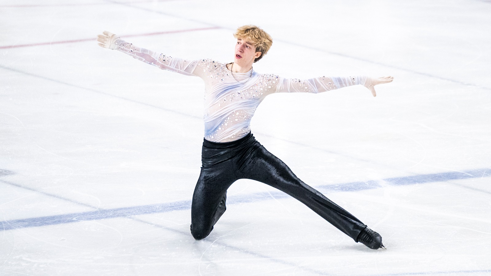Lucius Kazanecki kneels on the ice with both arms extended outward. He is a young man with blonde hair wearing a long sleeve white and light blue top with jewel detailing and shiny black pants. 