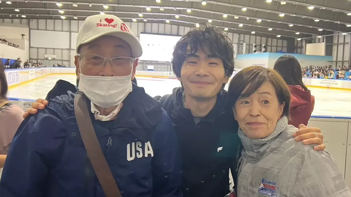 Goku stands in the middle of his grandpa (left) and grandma (right) at the Challenger Series event in Japan. The rink is behind them. his grandpa wears a navy jacket that says USA on the right chest and his grandma is in a grey U.s. Figure Skating jacket. Goku wears all black and they all smile. His grandpa is wearing a mask. 