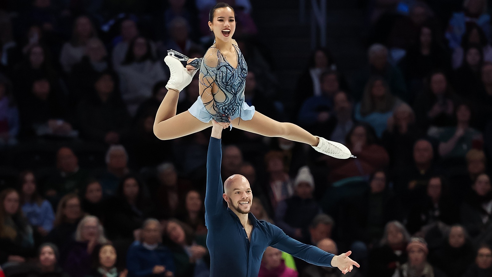 Ellie Kam and Danny O'Shea perform a lift with Danny holding Ellie up in the air, both smiling. Ellie is a young woman wearing a light blue skating costume and Danny is a man who is bald wearing a dark teal long sleeve top