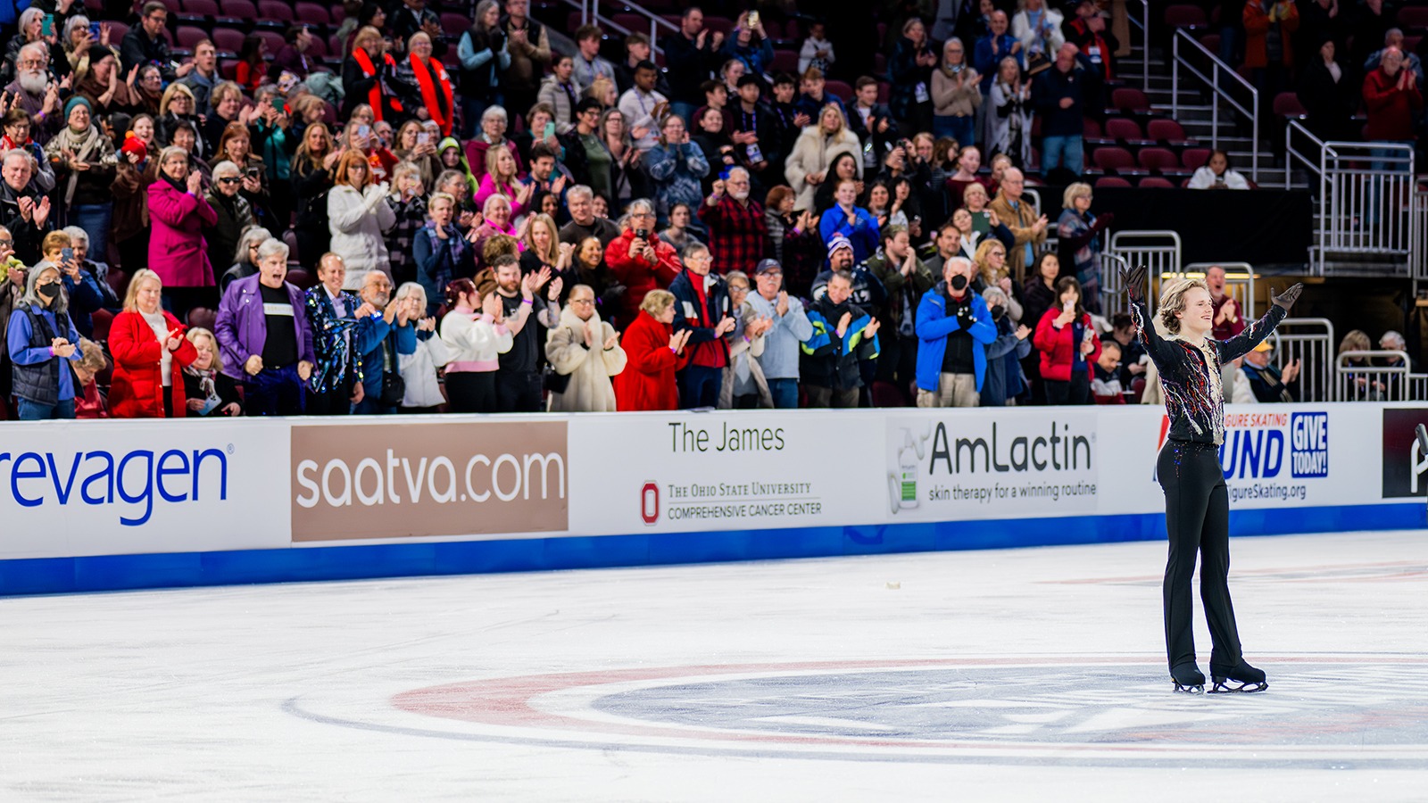 Ilia Malinin salutes the crowd with his arms outstretched and a smile on his face as people in the stands cheer. Ilia is a young man with short blonde hair wearing a black top with red and white stripes and black pants. 