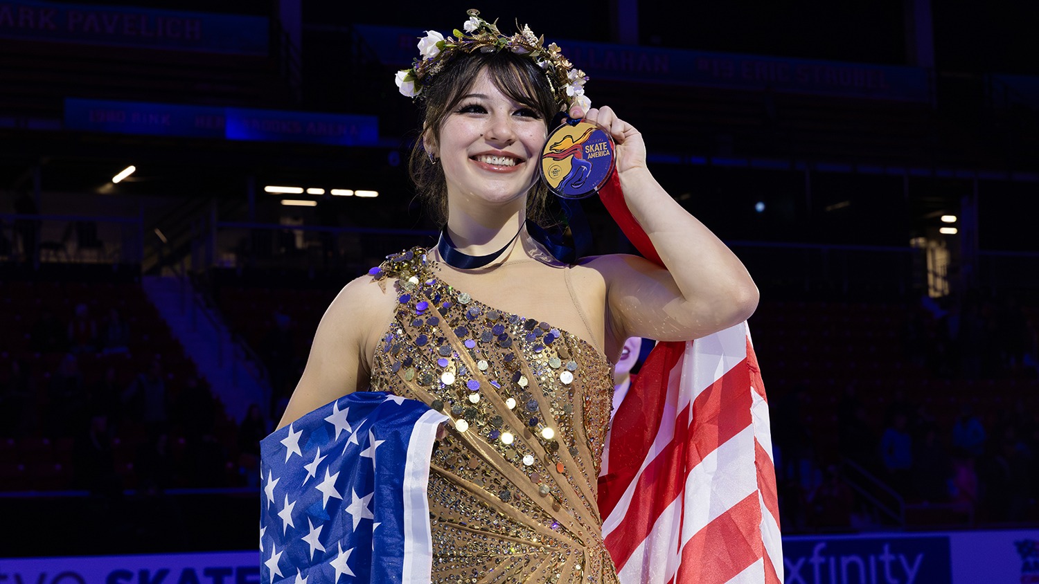 Alysa Liu, in a gold dress with gold sequins, poses with her medal during the medal ceremony at 2025 Saatva Skate America. She holds the medal to her cheek and smiles, a flower crown on her head. The U.S. flag is draped around her.