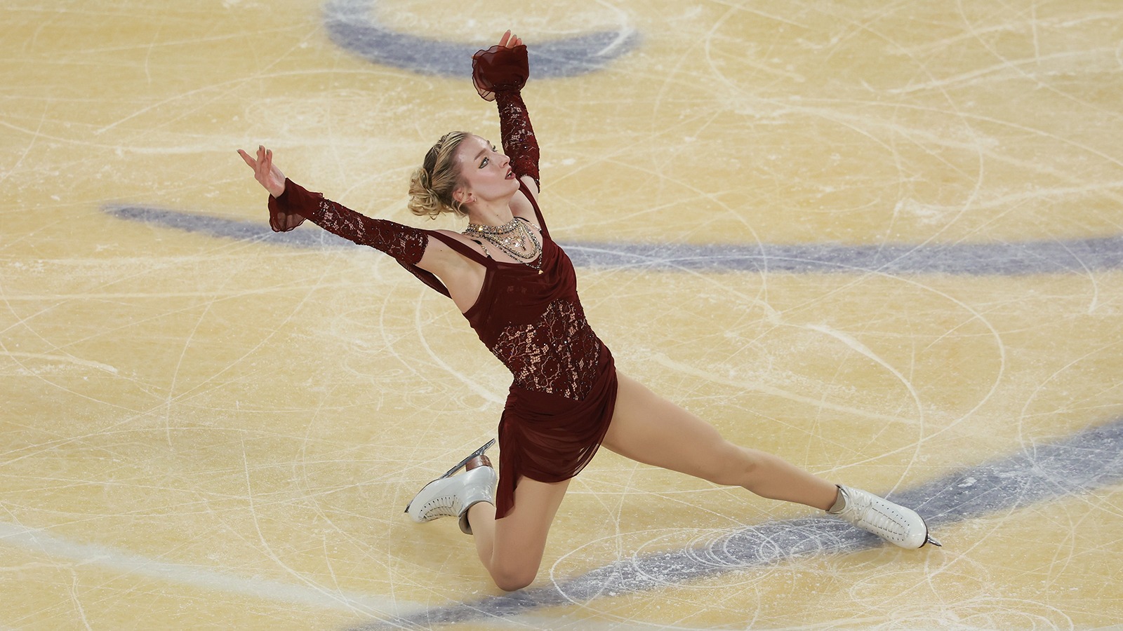 Amber Glenn kneels on the ice, looking up and with both arms raised above her head. Amber is a young woman with blonde hair tied back in a bun wearing a maroon skating costume with several chunky gold necklaces around her neck.